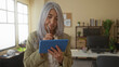 © Krakenimages.com - Woman with grey hair working on tablet in modern office setting with plants and shelves in background, reflecting focus and productivity.