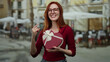 © Krakenimages.com - Woman smiling with gift on terrace, wearing glasses and red shirt, expressing joy outdoors at city restaurant.