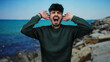 © Krakenimages.com - Man expressing frustration on rocky seaside with ocean waves under clear blue sky, wearing green sweater, hands covering ears, showcasing intense emotion and dramatic scenery.