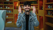 © Krakenimages.com - Young man smiling in a library setting with bookshelves behind, wearing a denim jacket, representing hispanic culture and adulthood indoors.