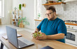© Studio Romantic - Overweight obese man eating salad in home kitchen. By laptop, he chooses healthy greens and a diet lunch, supporting weight goals during remote work. Nutrition and dieting concept.
