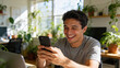© TimosBlickfang - Smiling young man using smartphone at home office filled with indoor plants, modern remote work and digital communication concept