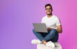 © Prostock-studio - Young Arab man in casual wear working online, sitting cross legged and using laptop in neon light, full length. Millennial Eastern guy surfing internet on portable pc, copy space