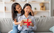 © Prostock-studio - Playful asian girl making surprise for her happy mother, closing her eyes and giving gift box while sitting on couch at home interior, copy space