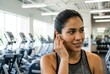 © Stitch - A smiling, sweaty Hispanic woman in a gym puts a wireless earbud in her ear, getting ready to listen to music during her workout.