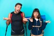 © luismolinero - Young mountaineer couple with a big backpack isolated on blue background having doubts while raising hands and shoulders