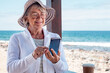 © luciano - Smiling senior woman wearing hat and eyeglasses standing at the beach using mobile phone typing message, horizon over sea and blue sky