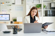 © wattana - Focused professional businesswoman working on a laptop while reviewing documents on a clipboard.