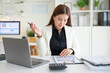 © wattana - Focused professional businesswoman working on a laptop while reviewing documents on a clipboard.