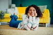 © deagreez - Bright happy young woman with curly hair lies on a rug in a colorful striped sweater at home on a sunny day smiling at the camera