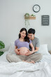 © Kawee - Asian husband and pregnant wife sit together on cozy bed in bedroom.
