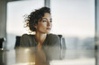 © EricMiguel - Pensive businesswoman with curly hair and hoop earring seated at a reflective glass conference table, gazing toward sunlit city window