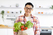 © sofiko14 - An Asian man smiles while holding a bag of fresh produce in his kitchen