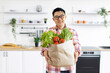 © sofiko14 - An Asian man smiles while holding a paper bag filled with fresh produce in his bright, modern kitchen