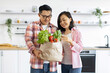 © sofiko14 - An Asian couple smiles while holding a bag of fresh groceries in their modern kitchen