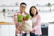 © sofiko14 - An Asian couple smiles while holding a bag of fresh produce in their kitchen