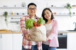 © sofiko14 - An Asian couple smiles while holding a bag of fresh produce in their bright, modern kitchen