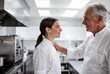 © zhou yan - Senior male chef in clean white apron places encouraging hand on shoulder of young female apprentice in bright modern professional kitchen with natural diffused lighting and shallow depth of field. Hi