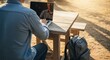 © Lena - Man working remotely on laptop at outdoor picnic table with backpack
