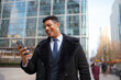 © Videophilia - A black man smiles as he looks at his phone while standing in an urban area. Tall buildings surround him, and people walk by in the background. It is a clear day.