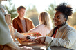 © Seventyfour - Young adult Black woman paying with credit card using wireless terminal at outdoor market stall, smiling at vendor while two Caucasian young adults talking in background