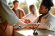 © Seventyfour - Black young adult woman making contactless payment at outdoor market counter while Caucasian vendor holding payment terminal, two young adults standing in background near produce