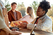 © Seventyfour - Young Black woman holding wooden basket of strawberries, preparing to pay with card at outdoor market counter, interacting with Caucasian man vendor while two young adults stand in background