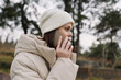 © SHOTPRIME STUDIO - Woman in a quilted beige coat and white beanie talks on a mobile phone outdoors, winter scenery with bare trees under gray skies, creating a calm candid moment. The scene emphasizes street fashion,