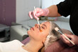 © Vagengeim - A cosmetology facial treatment is performed using a brush applicator on a woman lying on a treatment bed, with protective gloves and disposable cap visible in a professional skincare environment. Jet