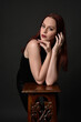 © faestock - close up portrait of beautiful female model with long red hair and black shirt, gestural hand and arm poses, leaning on wooden plinth. classic studio portrait on dark background.