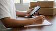 © Azizur - Asian Entrepreneur Managing Online Store Orders, Using a Smartphone and Writing Shipping Details on a Notepad with Parcels