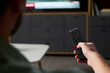 © pressmaster - Man holding television remote control, pointing toward flat screen TV with news broadcast visible on screen, sitting in modern living room environment