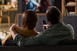 © pressmaster - Caucasian young adult man and Caucasian young adult woman sitting on sofa watching television together, man holding remote control, couple relaxing at home, backs facing camera