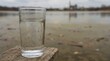 © K4VEE - A clear glass sits on a wooden dock edge with a muted, murky water scene behind, conveying clean water access and environmental concern with generous background copy space.