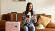 © Home-stock - Happy lady using cellphone and holding passport, sitting with packed suitcase and ordering taxi, sitting on sofa at home. Tourism and technology