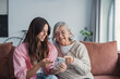 © Daniel - A young woman embraces her elderly mother as they sit on the sofa at home, smiling and looking at a phone together while watching content or shopping online in a relaxed family moment
