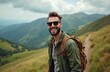 © Pete - Young man with backpack smiles on mountain path. He wears sunglasses and a green jacket while hiking. Green hills and cloudy sky in background.