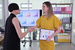 © H_Ko - Two businesswomen share a handshake, showcasing teamwork and accomplishment. Bright charts in the background illustrate their recent success in a contemporary office environment.