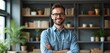 © Viktor - Smiling man with glasses wearing denim shirt stands arms crossed in a modern office with bookshelves. He looks friendly and professional, ready for business. Positive vibes.