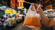 © weerasak - Thai iced tea in plastic bag with straw, held by hand at vibrant night market, colorful lights and street food stalls in background