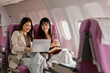 © kamiphotos - Two women sit side by side on an airplane, reviewing documents and working on a laptop together, showing teamwork, shared focus, and a calm working atmosphere during air travel.