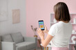 © Pixel-Shot - Beautiful young woman using mobile phone with smart home security system control panel in room, back view