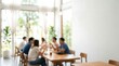 © CuteBee - A blurred photograph captures diverse individuals conversing around a wooden table in a modern, bright cafe, suggesting a relaxed informal meeting.