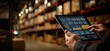 © Akbar - Hand holding tablet displaying warehouse analytics near shelves of boxes