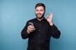 © bodnarphoto - Young man smiles while holding smartphone in front of blue background during day time