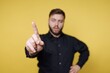 © bodnarphoto - Man holding up one finger against yellow background while wearing a black shirt and having a serious expression on his face