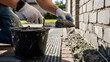 © Mahmuda - Construction worker applying cement mortar to brick wall foundation | Professional mason using trowel for house exterior repair | Close-up of gloved hands mixing concrete for building job