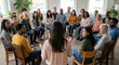 © Jana - A female facilitator gestures while addressing a large, diverse group of adults seated in a circle during a support group meeting or therapy session.