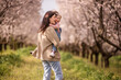 © farmuty - Mother carries smiling daughter through blooming almond orchard, sharing a joyful spring moment of bonding and warmth among green grass and sunlit trees