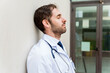 © StockMe - Overworked male doctor with a uniform and stethoscope leaning against a wall, resting with his eyes closed, showing fatigue
